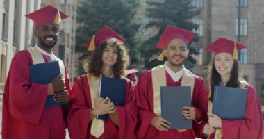 Mezuniyet mantolarındaki kadın ve erkeklerin yavaş çekimde portresi ve üniversite kampüsünde ellerinde diplomalarla duran havan tahtaları. Yüksek öğrenim ve hırslı gençlik kavramı.