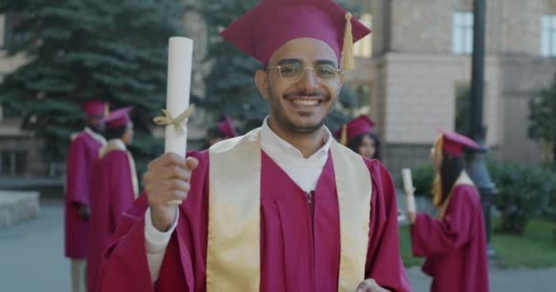 快乐的中东男人挥动着文凭 指着站在大学旁边笑着的卷轴慢动作肖像 教育和成绩概念 — 图库视频影像