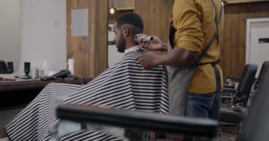 Berber dükkanında modern saç stili yaratan yetenekli bir berber müşteriyi makineyle tıraş ederken yan görüş. Moda ve güzellik salonu konsepti.