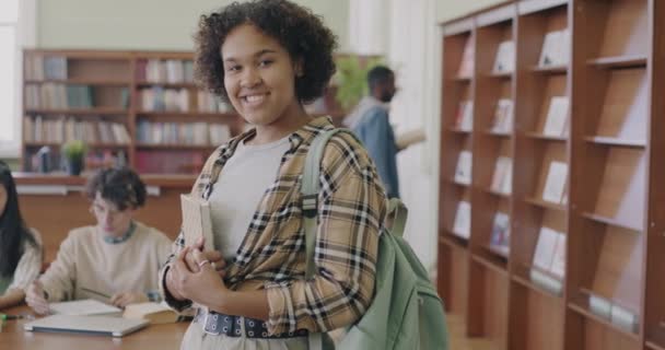 スローモーションポートレートの陽気なアフリカ系アメリカ人女性学生立って図書館で本やバックパック笑顔カメラを見て — ストック動画
