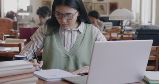 快乐的亚洲女学生在大学图书馆使用笔记本电脑打字 浏览笔记和在笔记本上写作 学术教育和技术概念 — 图库视频影像