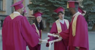 Çok ırklı mezunlar diplomalarla el ele tutuşup çığlık çığlığa gülüyorlar ve üniversite kampüsünde eğleniyorlar. Eğitim ve eğlence konsepti.