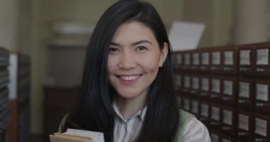 Lise kütüphanesinde elinde kameraya bakarak gülümseyen kitaplar tutan neşeli Asyalı kız öğrencinin yavaş çekimde portresi. Halk ve eğitim kavramı.