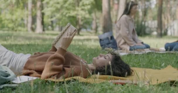 一个无忧无虑的年轻女子躺在草坪上看书的画像 在公园里欣赏着现代文学 闲暇时间概念中的人和业余爱好 — 图库视频影像