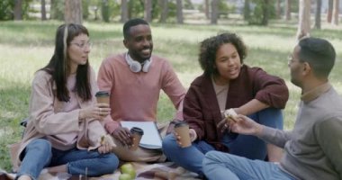 Mutlu genç arkadaşlar parkta piknik yapmaktan, sandviç yemekten ve kahve bardakları tokuşturmaktan keyif alıyorlar. Modern yaşam tarzı ve arkadaşlık kavramı.