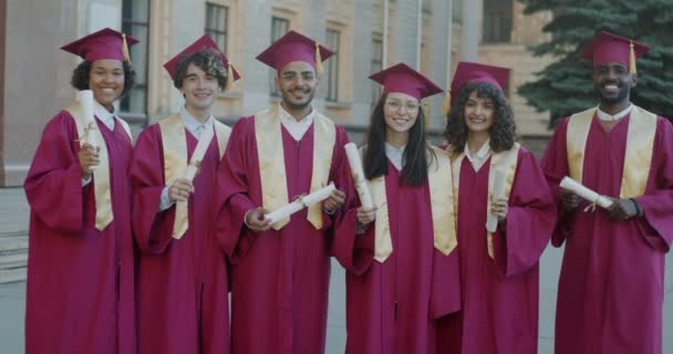 Różnorodna Grupa Studentów Stojąca Kampusie Uniwersyteckim Sukniach Kapeluszach Uśmiechniętymi Dyplomami — Wideo stockowe