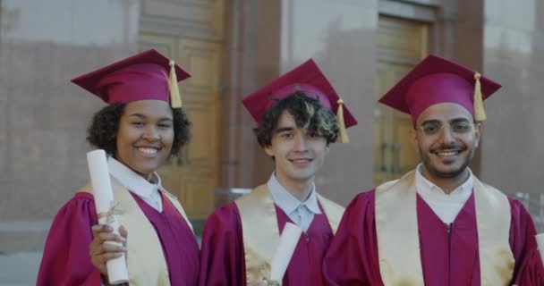 Multi-ethnic group of people in gowns and graduation hats standing on campus holding diplomas smiling looking at camera. Youth and higher education concept.