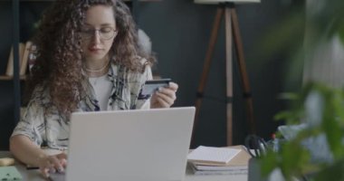 Young lady paying with bank card online using laptop typing holding plastic card indoors at desk at home. People and banking concept.