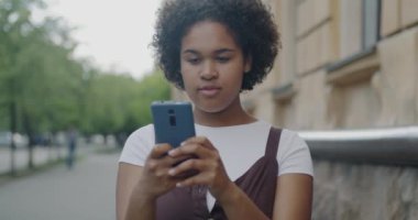 Portrait of happy African American woman using smartphone touching screen standing in city street. Modern gadgets and technology concept.