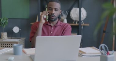 Cep telefonuyla konuşan ve ofisteki laptop ekranına bakan genç iş adamının portresi. İş iletişimi ve iş kavramı.