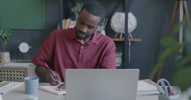 アフリカ系アメリカ人のビジネスマンは ラップトップとワイヤレスイヤホンでオンラインビデオ通話を行い オフィスで執筆しています ビジネスとコミュニケーションのコンセプト — ストック動画