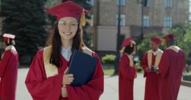 Kampüste elinde diplomayla duran hırslı genç bir bayan öğrencinin ağır çekimde portresi. Halk ve yüksek öğrenim kavramı.