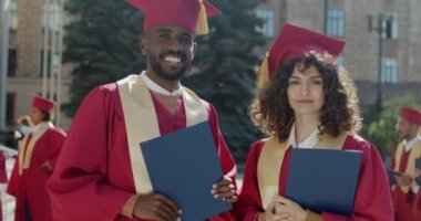 Kampüste diploma taşıyan havan ve manto takan kadın ve erkek öğrencilerin yavaş çekimde portresi. Gençlik ve eğitim kavramı.