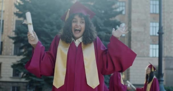 大学校园里戴着毕业帽 身穿长袍 笑着挥动文凭的女孩的慢动作肖像 情感和教育概念 — 图库视频影像