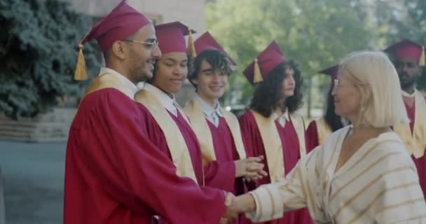 成熟的女大学教授与年轻人握手 拥抱穿着礼服和帽子的毕业生 毕业典礼和教育观念 — 图库视频影像