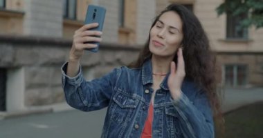 Şehrin arka planında akıllı telefon kamerasıyla selfie çeken güzel bir kadının yavaş çekim portresi. Kadehler ve fotoğrafçılık kavramı.