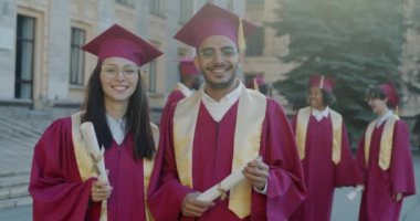 Beyaz kadın ve Arap adam, mezuniyet kepleri ve cübbeleri takmış, elinde diploma tomarları ve gülümsemeyle kampüste duruyorlar. Halk ve eğitim kavramı.