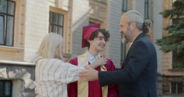 家长们在校园里拥抱着穿着毕业礼服和砂浆板的儿子 祝贺他们 高等教育和家庭关系概念 — 图库视频影像