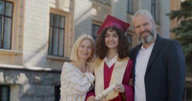 Sabahlık ve şapkayla mezun olan genç bir kadının yavaş çekimde portresi ve kampüste anne ve babasının gururu. Gülümseyen insanlar.