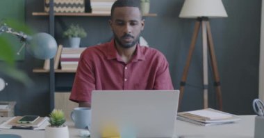 Genç Afrikalı Amerikalı erkek öğrencinin portresi dizüstü bilgisayarla çalışıyor ve evdeki masada gülümsüyor.