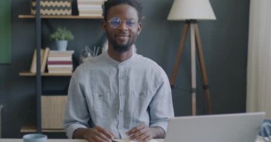 Modern stüdyo dairesindeki masasında oturmuş kameraya gülümseyen neşeli Afrikalı Amerikalı adamın yavaş çekimde portresi. İnsanlar ve duygu ifade kavramı.