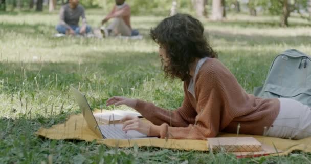 公園で試験のために勉強している若い女性は ラップトップと一緒に作業し 芝生の毛布の上に横たわる教科書を読んでいます 教育と自然のコンセプト — ストック動画