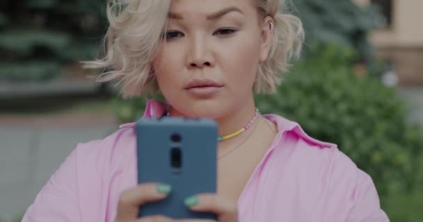 スマートフォンのタッチスクリーンでブロンドの髪メッセージングと街の通りで微笑むかわいいアジアの少女 現代のガジェットと人々のコンセプト — ストック動画
