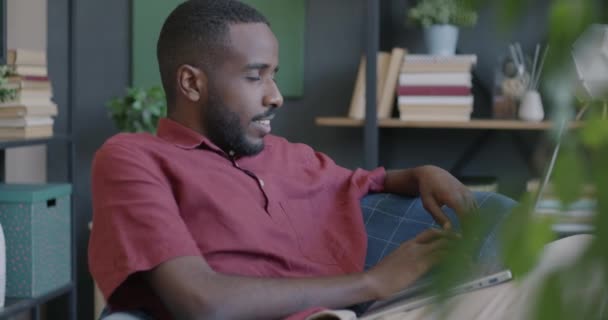 自宅で快適なソファーに座っているラップトップタイピングを使用して幸せなアフリカ系アメリカ人男性 現代のテクノロジーとソーシャルメディアのユーザーコンセプト — ストック動画