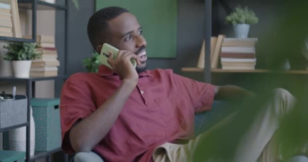 ハッピーアフリカ系アメリカ人男性は 昼間のアパートで椅子に座っている携帯電話で話しています スマートフォンコミュニケーションとホームコンセプト — ストック動画