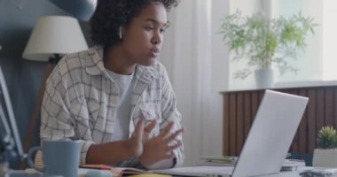 Afrikalı Amerikalı öğrenci çevrimiçi video görüşmesi sırasında çevrimiçi konuşuyor ve evdeki masada deftere yazıyor. Uzaktan eğitim ve e-öğrenme kavramı.
