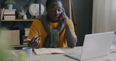 Serbest çalışan Afrikalı Amerikalı adam cep telefonuyla konuşuyor evdeki laptop ekranına bakıyor. İletişim ve serbest meslek kavramı.