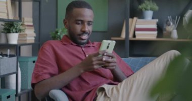 Sosyal medyada akıllı telefon mesajları kullanan neşeli bir öğrenci. Oturduğu koltukta oturuyor. Modern teknoloji ve çevrimiçi iletişim kavramı.