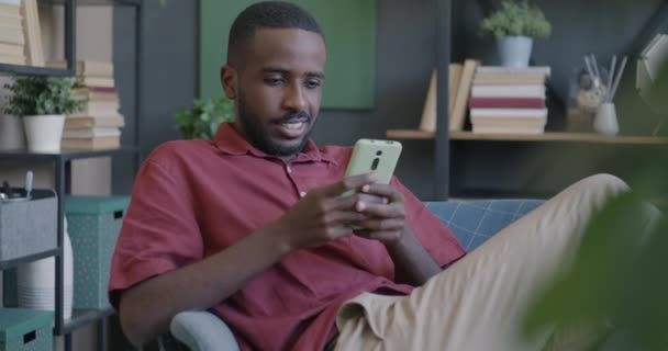 アパートのアームチェアに座っているソーシャルメディアでスマートフォンテキストを使用して楽しい学生 現代技術とオンラインコミュニケーションのコンセプト — ストック動画