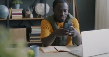 Genç Afro-Amerikan erkek öğrenci akıllı telefon uygulamasını evde masada otururken kullanıyor. Modern teknoloji ve çevrimiçi içerik kavramı.