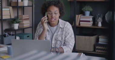 Genç Afrikalı Amerikalı kadın müşteriyle cep telefonuyla konuşuyor ve ofis bilgisayarını kullanıyor. İletişim ve mesleki işgal kavramı.
