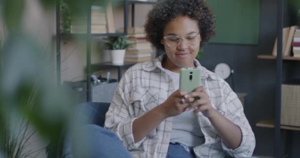 无忧无虑的非洲裔美国女人在家里的扶手椅上用智能手机浏览放松的肖像 现代小玩艺儿和青少年概念 — 图库视频影像