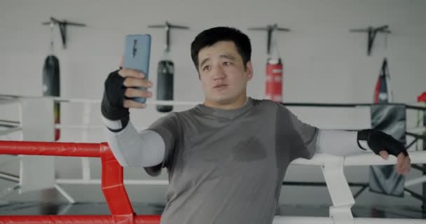 陽気なアジアのスポーツマンは ボクシング機器を示すスマートフォンカメラを使用してジムからオンラインビデオ通話を作ります スポーツとコミュニケーションのコンセプト — ストック動画