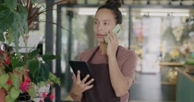 Çiçekçi dükkanı sahibi cep telefonuyla konuşuyor ve iş geliştirme hakkında tablet kullanıyor. Arka planda görünen güzel yeşil saksı bitkileri.
