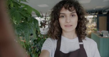 Önlüklü neşeli genç bir kadın internette konuşuyor ve çiçekçide yeşil bitkileri işaret ederek gülüyor. İş ve iletişim kavramı.