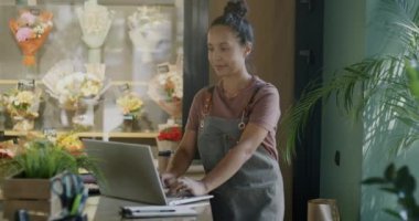 Genç bir çiçekçi kadın dizüstü bilgisayar kullanırken bayan çalışanlar dükkanda bitkilerle dolu kutular getiriyor. Perakende ve ticaret kavramı.