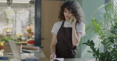 Çiçekçide dizüstü bilgisayarla çalışan ve cep telefonuyla konuşan neşeli bayan girişimci. İş iletişimi ve teknoloji kavramı.