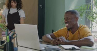 Kadın arka planda çalışırken, Afrikalı Amerikalı bir adam modern çiçekçi dükkanında dizüstü bilgisayarla çevrimiçi video görüşmesi yapıyor. İletişim ve iş konsepti.