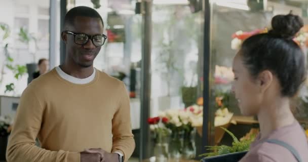 アフリカ系アメリカ人男性が花を買って 店のセールスウーマンから花束を取ってスマートフォンで支払う 金融コンセプト — ストック動画