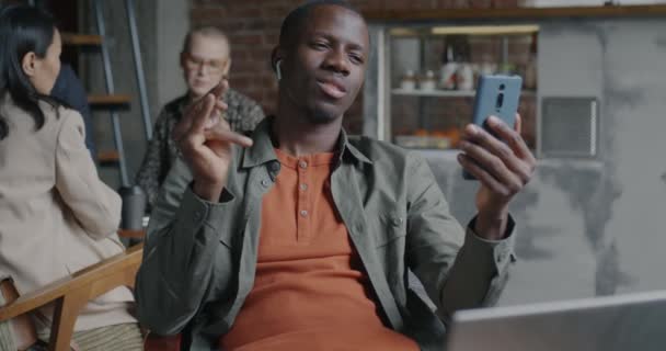 カフェからスマートフォンとワイヤレスイヤホンでオンラインビデオ通話をするケアフリーのアフリカ系アメリカ人男性 バーチャルミーティングとコミュニケーションコンセプト — ストック動画