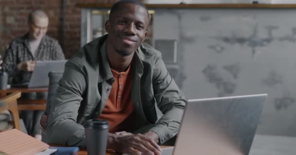 自信を持ったアフリカ系アメリカ人の男性が コワーキングオフィスのノートパソコンでテーブルに座って微笑んでいます ビジネスと人々のコンセプト — ストック動画