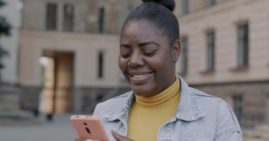 Neşeli Afro-Amerikan kadın akıllı telefon kullanarak açık havada gülümsüyor. Modern aygıtlar ve iletişim konsepti.