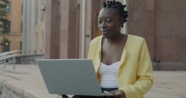 Afro-Amerikalı neşeli kız öğrenci internette bilgisayarla video görüşmesi yapıyor. Şehir caddesinde oturuyor. İletişim ve insan kavramı.