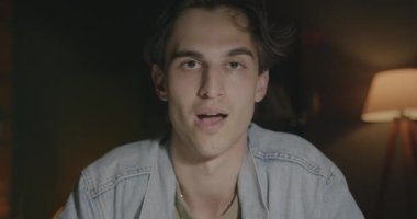 Çevrimiçi video konuşması yapan neşeli genç bir adamın portresi el sallayarak karanlık arka planda kameraya bakıyor. İletişim ve konferans konsepti.