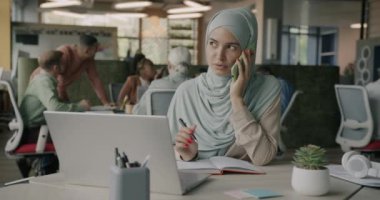 Dost canlısı Müslüman kadın cep telefonuyla müşteriye konuşuyor ve yaratıcı açık uzay ofisinde laptopunu kullanıyor. İş iletişimi ve çalışanlar kavramı.