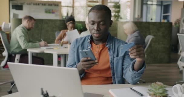 クリエイティブオフィスで働くスマートフォンとクレジットカードでオンライン決済をするアフリカ系アメリカ人男性 ファイナンスと現代技術のコンセプト — ストック動画